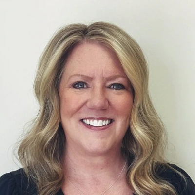 Person with shoulder-length, wavy blonde hair wearing a dark top and a necklace